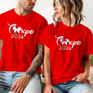 Puede incluir: Dos camisetas rojas con texto blanco "Tokyo 2026" en una fuente estilizada. Una camiseta tiene un dise&ntilde;o de cabeza de Mickey Mouse, la otra un dise&ntilde;o de cabeza de Minnie Mouse. Ambas camisetas son de manga corta.