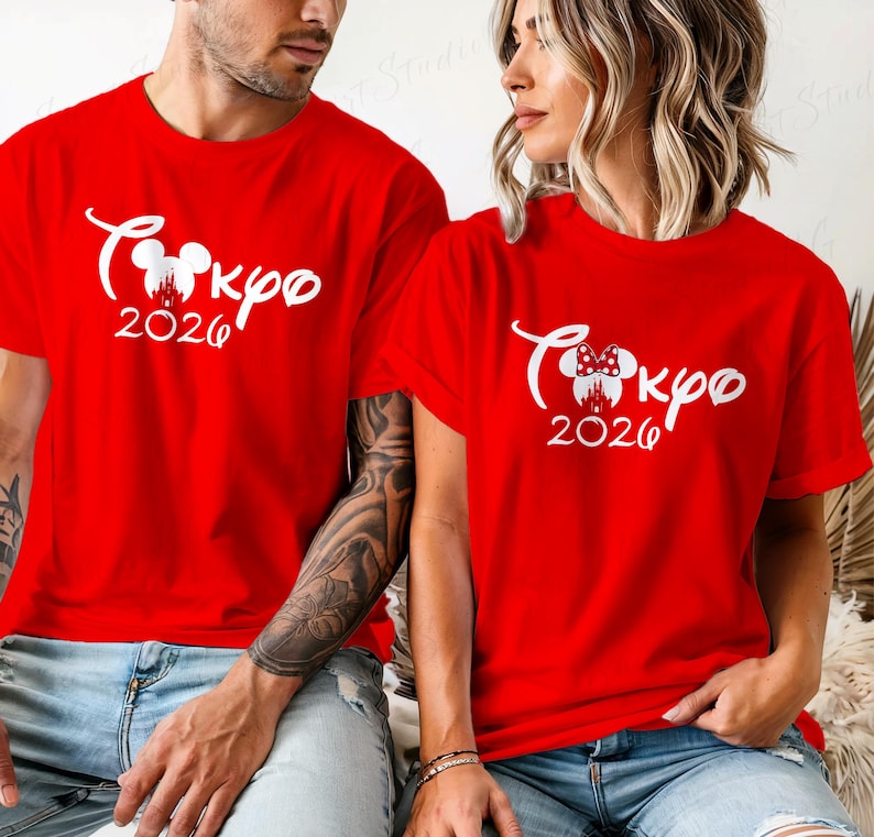 Puede incluir: Dos camisetas rojas con texto blanco "Tokyo 2026" en una fuente estilizada. Una camiseta tiene un dise&ntilde;o de cabeza de Mickey Mouse, la otra un dise&ntilde;o de cabeza de Minnie Mouse. Ambas camisetas son de manga corta.