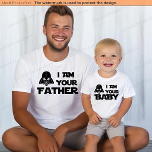 Pode incluir: Camisetas brancas combinando com texto preto e gráficos de Darth Vader. Uma camisa diz "I AM YOUR FATHER" e a outra "I AM YOUR BABY". As camisas são usadas por um pai e seu filho.