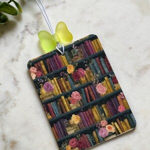 May include: A rectangular hanging air freshener featuring a printed design of a bookshelf filled with colorful books and interspersed with various pink, yellow, and orange floral blooms. A translucent yellow butterfly bead sits above.
