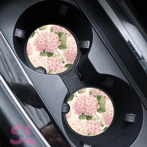May include: Two car coasters with a floral hydrangea design. The coasters are round and feature a cream background with pink hydrangea blooms and green leaves. They are placed in a black car cup holder.
