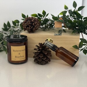 Może przedstawiać: Martwa natura z brązowym szklanym słoikiem na świece z czarną pokrywką i napisem "Butterscotch & Bourbon". Obecny jest również brązowy flakon z rozpylaczem, mała szklana butelka, szyszki i zielone liście. Kompozycja jest prezentowana na jasnym tle.