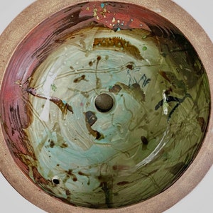 May include: A ceramic bowl with a unique, abstract design. The interior features a blend of green, blue, and brown hues with dark, organic-shaped patterns. The rim is a reddish-brown color, and the outer edge is a lighter tan. The bowl has a central drain hole.