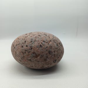Pode incluir: Uma pedra arredondada, castanho-avermelhada, com uma superfície texturizada. A pedra tem manchas escuras e um aspeto liso e polido. A pedra natural é um objeto decorativo.