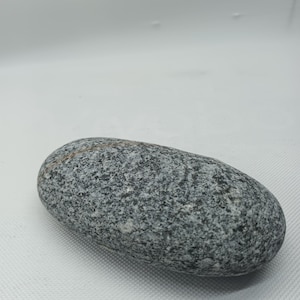 Könnte beinhalten: Ein glatter Flussstein mit grau-schwarzer Sprenkelung. Der längliche Stein hat eine abgerundete Form und ein poliertes Aussehen. Der Stein ist ein Naturprodukt mit Farb- und Texturvariationen. Der Stein hat eine neutrale Farbe und kann zur Dekoration verwendet werden.
