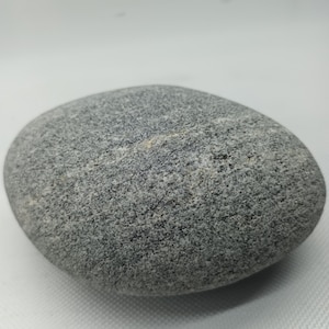 Peut inclure: Une pierre grise lisse de forme arrondie. La surface de la roche est texturée avec de petits détails granulaires. La couleur est un mélange de gris clair et foncé, avec de subtiles variations. La pierre semble naturelle et non polie.