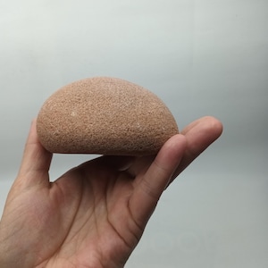 Könnte beinhalten: Ein glatter, rotbrauner, halbmondförmiger Stein. Der Stein hat eine strukturierte Oberfläche und wird in einer Hand vor einem schlichten weißen Hintergrund gehalten. Es handelt sich wahrscheinlich um einen Naturstein.