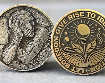 May include: Two antique-style metal coins. One coin features a detailed relief of a person holding their head. The other coin has the text "LET BOREDOM GIVE RISE TO IDEAS AND ACTION" around a sun, plant, and moon design.