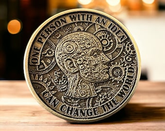 May include: A bronze-colored coin with the text "ONE PERSON WITH AN IDEA CAN CHANGE THE WORLD" encircling a detailed profile of a human head. The head is intricately designed with gears and mechanical elements. The coin is on a wooden surface.