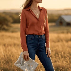 Könnte beinhalten: Eine Frau trägt einen beigefarbenen Cowboyhut, ein rostfarbenes Hemd und dunkelblaue Jeans. Sie hält eine Clutch mit einem grau-goldenen Muster und einer Quaste. Der Hintergrund ist ein Feld mit einer Scheune.