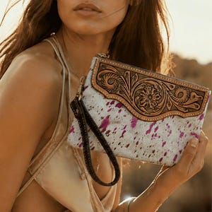 Könnte beinhalten: Eine Clutch mit weißen und rosa Flecken und einem braunen Leder-Handgelenksriemen. Die Clutch hat eine braune Lederklappe mit floralen Prägungen. Die Frau im Bild trägt ein goldenes Kleid und silberne Armbänder.