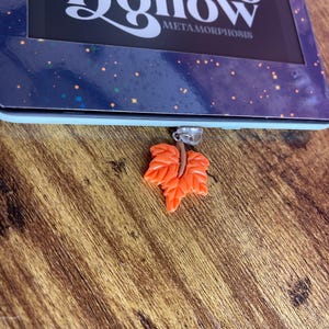 May include: A close-up of an orange leaf-shaped bookmark with a silver ring. The bookmark is attached to a book with the title "Grove Hollow Metamorphosis".