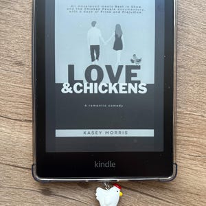 Puede incluir: Un lector de libros electrónicos Kindle negro que muestra la portada del libro "Love & Chickens" de Kasey Morris. La portada presenta una pareja tomados de la mano y una ilustración de un pollo. Un dije de pollo blanco con cresta roja y huevo naranja cuelga del dispositivo.