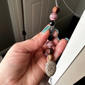May include: A beaded charm with a variety of colors and textures, including black, brown, pink, and white. The charm features a small metal flower charm and a large oval bead with a cracked pattern.