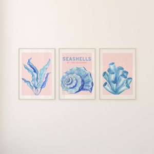 Könnte beinhalten: Drei gerahmte Drucke mit rosa Hintergrund und blauen Aquarell-Illustrationen von Muscheln, Algen und Korallen. Der mittlere Druck trägt den Text "Seashells by the Seashore".