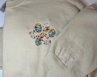 May include: A beige sweatshirt with a floral paw print embroidery on the chest. The embroidery features a variety of colorful flowers. The name "Max" is embroidered on the cuff of the sleeve.