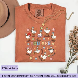 May include: A burnt orange Comfort Colors t-shirt featuring a graphic of cartoon ducks with positive affirmations like "Brave," "Kind," and "Strong." The shirt includes the text "YOU ARE" and is adorned with floral and star accents. A gold necklace with a star pendant is draped on the shirt.