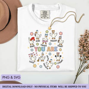 May include: White Comfort Colors t-shirt with a crew neck, featuring a colorful graphic of cartoon geese with positive affirmations. The words "YOU ARE" are in the center, surrounded by words like "Brave", "Kind", and "Smart". A gold star necklace is draped on the shirt.
