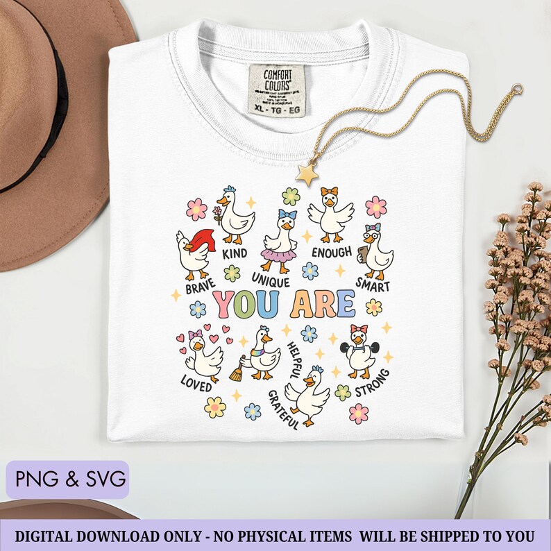 May include: White Comfort Colors t-shirt with a crew neck, featuring a colorful graphic of cartoon geese with positive affirmations. The words "YOU ARE" are in the center, surrounded by words like "Brave", "Kind", and "Smart". A gold star necklace is draped on the shirt.