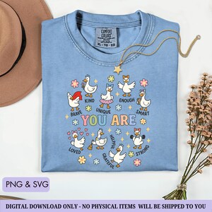 May include: A light blue Comfort Colors t-shirt with a graphic of cartoon ducks with positive affirmations. The shirt has the words "You Are" in colorful letters, surrounded by ducks labeled "Brave," "Kind," "Unique," "Enough," "Smart," "Loved," "Helpful," "Grateful," and "Strong."