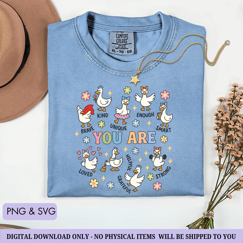 May include: A light blue Comfort Colors t-shirt with a graphic of cartoon ducks with positive affirmations. The shirt has the words "You Are" in colorful letters, surrounded by ducks labeled "Brave," "Kind," "Unique," "Enough," "Smart," "Loved," "Helpful," "Grateful," and "Strong."