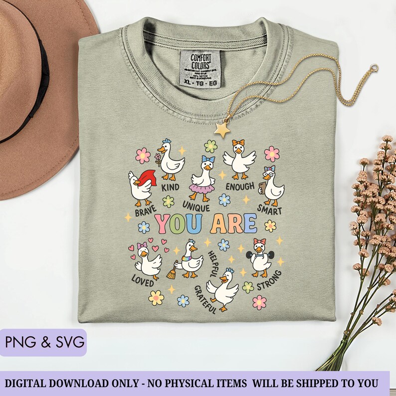 May include: Sage green Comfort Colors XL t-shirt with a graphic of cartoon ducks and the words "YOU ARE" surrounded by positive affirmations. A gold necklace with a star pendant is draped on the shirt. The text "PNG & SVG" is at the bottom.