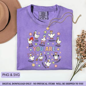 May include: A lilac Comfort Colors t-shirt with a graphic of cartoon ducks, each with a different accessory and word: Brave, Kind, Unique, Enough, Smart, Loved, Helpful, Grateful, and Strong. A gold necklace with a star pendant is draped on the shirt.