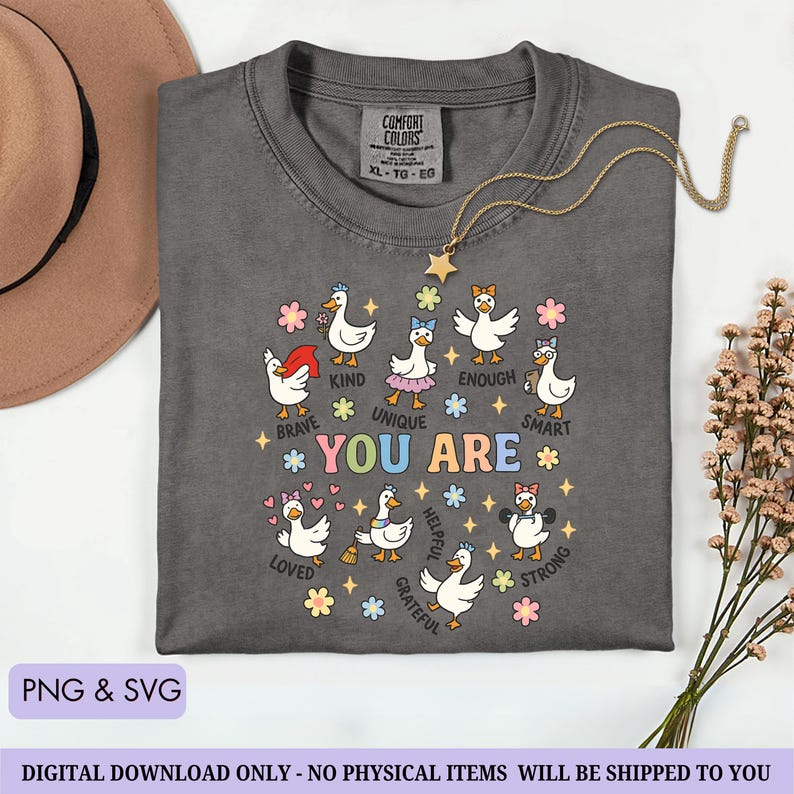 May include: Dark gray Comfort Colors t-shirt with a crew neck. The shirt features a colorful graphic of ducks with positive affirmations like "Brave," "Kind," and "Unique." A gold necklace with a star pendant is draped on the shirt. The words "PNG & SVG" are at the bottom.