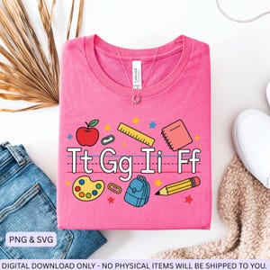 May include: A pink t-shirt with a school-themed design. The graphic includes an apple, ruler, notebook, paint palette, backpack, and pencil. The letters "Tt Gg Ii Ff" are displayed. The shirt is made of soft material.