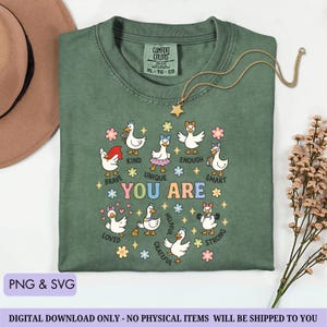 May include: Green Comfort Colors t-shirt with a graphic of cartoon ducks and the words "YOU ARE" surrounded by positive affirmations. A gold star necklace and a brown hat are also visible. The shirt is a size XL-TG-EG.