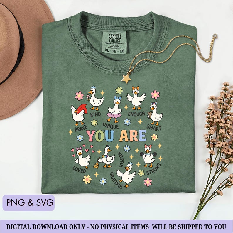 May include: Green Comfort Colors t-shirt with a graphic of cartoon ducks and the words "YOU ARE" surrounded by positive affirmations. A gold star necklace and a brown hat are also visible. The shirt is a size XL-TG-EG.