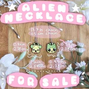 Op de afbeelding: Twee kettingen met een alien-thema met kettingen van 45 cm. Eén hanger is een groene hondachtige alien, de andere is een groene katachtige alien. De hangers zijn 2,38 cm breed. De tekst "ALIEN NECKLACE" en "FOR SALE" is roze.