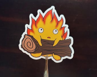 May include: A sticker featuring a cartoon fire spirit with a yellow body, orange and red flames, and wide eyes. The spirit is holding a brown log. The sticker has a white border and is on a dark background.