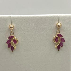 May include: A pair of gold drop earrings featuring a cluster of marquise-cut deep red gemstones arranged in a leaf-like pattern. Each earring is suspended from a small gold ball stud, showcasing a polished metal finish and vibrant red stones.