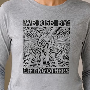 May include: A heather gray long-sleeve shirt featuring a black graphic print of multiple hands reaching toward a central hand above a sunburst design. The text "WE RISE BY LIFTING OTHERS" is printed in a bold, block font above and below the illustration.