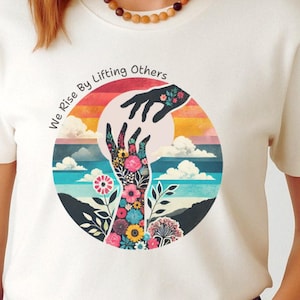 May include: Cream-colored t-shirt with a circular graphic featuring hands reaching towards each other. The graphic includes flowers, clouds, and the text "We Rise By Lifting Others."