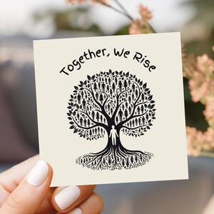 May include: A square card featuring the text "Together, We Rise" above a black tree illustration. The tree's branches and leaves are filled with small human figures. The card is held by a person with white nail polish.
