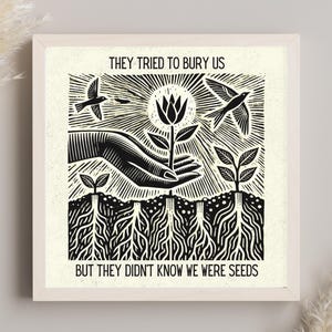 May include: A square framed print featuring a black and white linocut style illustration of a hand holding a flower with roots extending into the soil. Two birds fly above. The text reads: THEY TRIED TO BURY US BUT THEY DIDN'T KNOW WE WERE SEEDS.