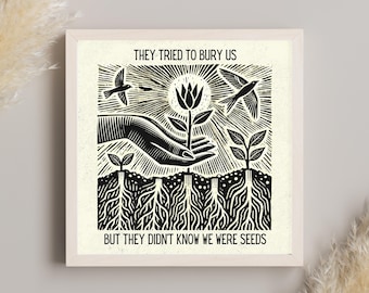 May include: Black and white art print in a light wood frame. The print features a hand holding a flower, with birds and the text "THEY TRIED TO BURY US BUT THEY DIDN'T KNOW WE WERE SEEDS". The artwork has a woodcut style.