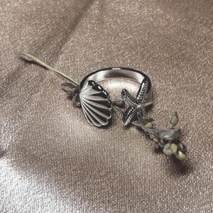 Op de afbeelding: Een zilveren ring met een schelp- en zeesterontwerp. De schelp heeft gedetailleerde lijnen en de zeester heeft kleine, donkere accenten. De ring wordt gepresenteerd op een getextureerd, lichtbruin oppervlak.