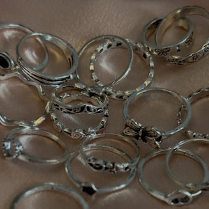 May include: A selection of silver rings featuring diverse designs, such as etched patterns, a bow, and a black stone. The rings are arranged on a soft, peach-coloured surface, presenting a delicate and elegant appearance.