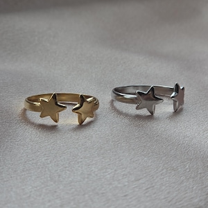 Puede incluir: Dos anillos abiertos, uno dorado y otro plateado, cada uno con dos adornos en forma de estrella. Los anillos se exhiben sobre una superficie suave de color neutro, mostrando su diseño simple pero elegante.