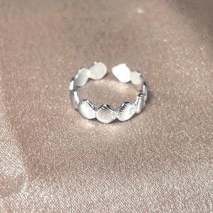 May include: A silver ring with a seashell design. The adjustable ring features a series of small, detailed shell shapes around the band. The ring is set against a soft, textured background.