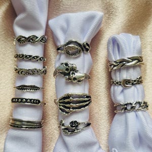 Könnte beinhalten: Eine Sammlung silberfarbener Ringe, präsentiert auf weißem Stoff. Die Ringe weisen verschiedene Designs auf, darunter geflochtene Bänder, eine Schlange, ein Schädel, eine Skeletthand, eine Rose und Kettenglieder. Der Hintergrund ist hellrosa.