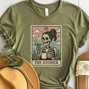 Puede incluir: Camiseta verde oliva con un diseño de carta del tarot que muestra un esqueleto fumando y sosteniendo una taza. La carta dice "III" y "THE STONER" en un estilo vintage. La camiseta se combina con un sombrero marrón y sandalias.