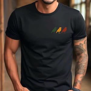 May include: A black short-sleeved t-shirt featuring a small graphic on the left chest area. The graphic consists of three stylized bird silhouettes arranged in a horizontal row, colored in muted green, yellow, and reddish-orange tones.