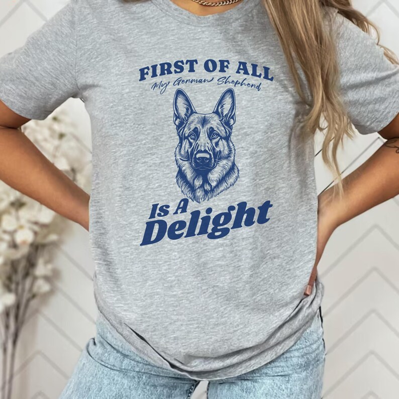 Pu&ograve; includere: Maglietta grigia con una grafica blu di un pastore tedesco e il testo "First of all, my German Shepherd is a Delight."