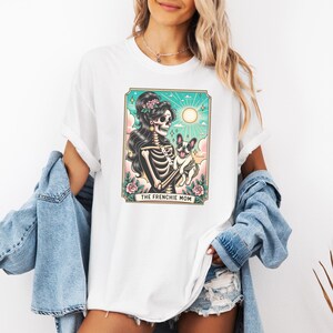 Pu&ograve; includere: Maglietta bianca con una grafica di uno scheletro che tiene un bulldog francese. La grafica &egrave; in stile vintage e ha il testo "The Frenchie Mom" in basso.