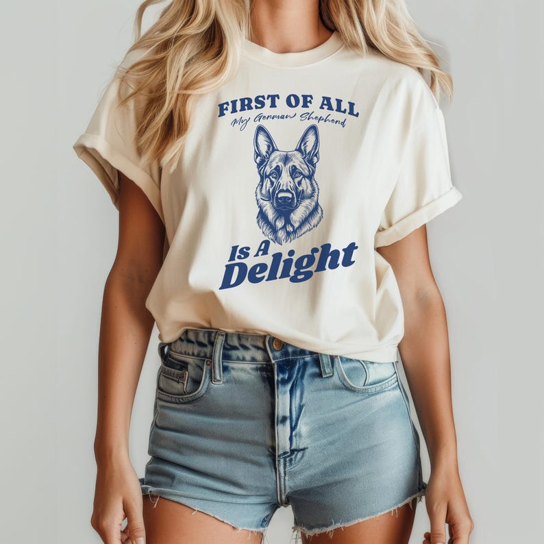 Pu&ograve; includere: Una maglietta bianca con una grafica blu di un pastore tedesco e il testo "First of all, my German Shepherd is a delight."