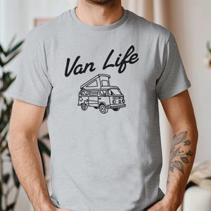 May include: A light gray t-shirt featuring the words "Van Life" in a black script above a black line drawing of a camper van. The van has a pop-up roof and is detailed with windows and wheels.